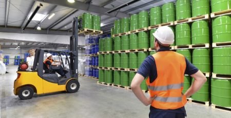 Warehouse worker in hi-vis vest overseeing forklift moving green drums, showing industrial radiant heating application