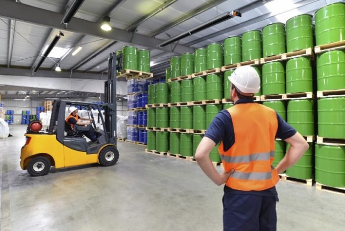 Warehouse worker in hi-vis vest overseeing forklift moving green drums, showing industrial radiant heating application