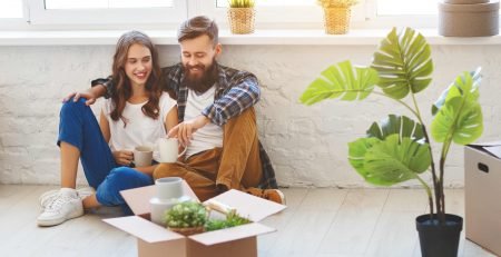 Happy couple sitting on warm floor of new home surrounded by moving boxes and plants