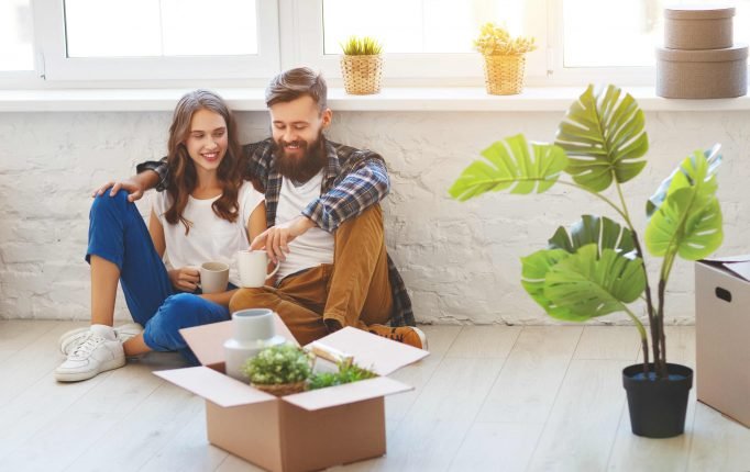 Happy couple sitting on warm floor of new home surrounded by moving boxes and plants