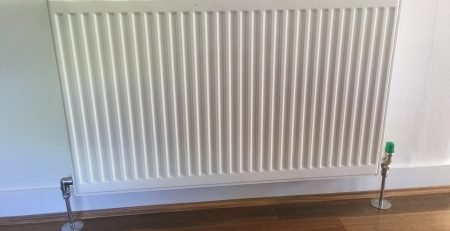 White hydronic panel radiator installed on wall above timber flooring with chrome pipe connections and thermostatic valve