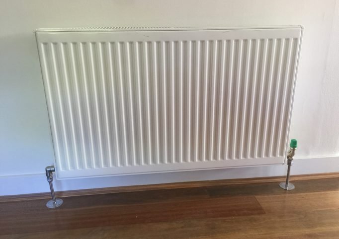 White hydronic panel radiator installed on wall above timber flooring with chrome pipe connections and thermostatic valve